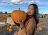 A woman holding a pumpkin and smiling in a pumpkin patch on a sunny day. She is wearing a brown jumper and has her hair down wearing a bold lipstick